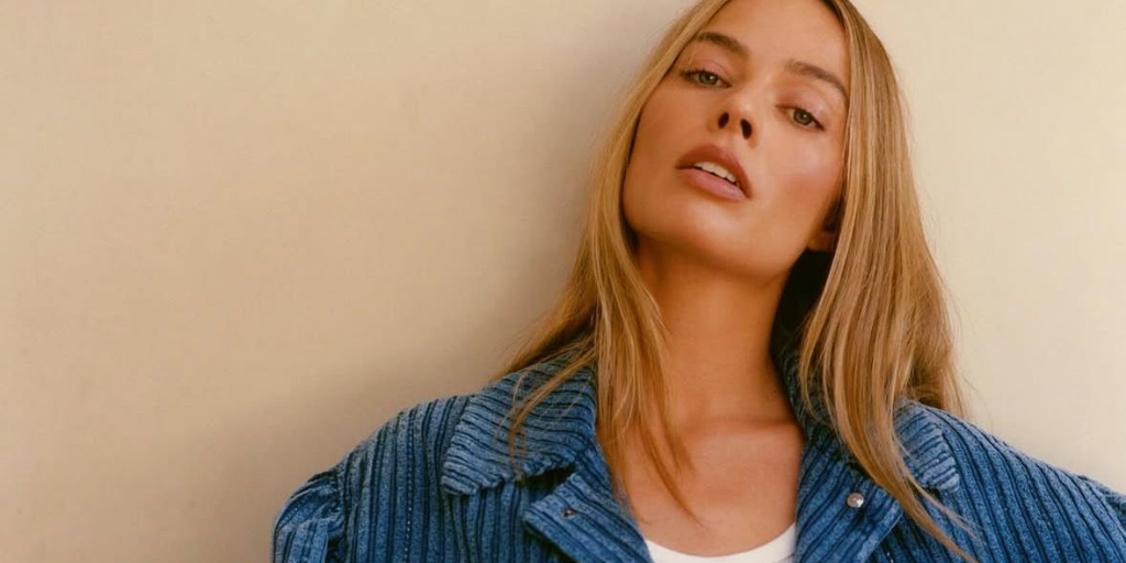 Margot Robbie leaning against a wall, wearing a blue corduroy jacket.