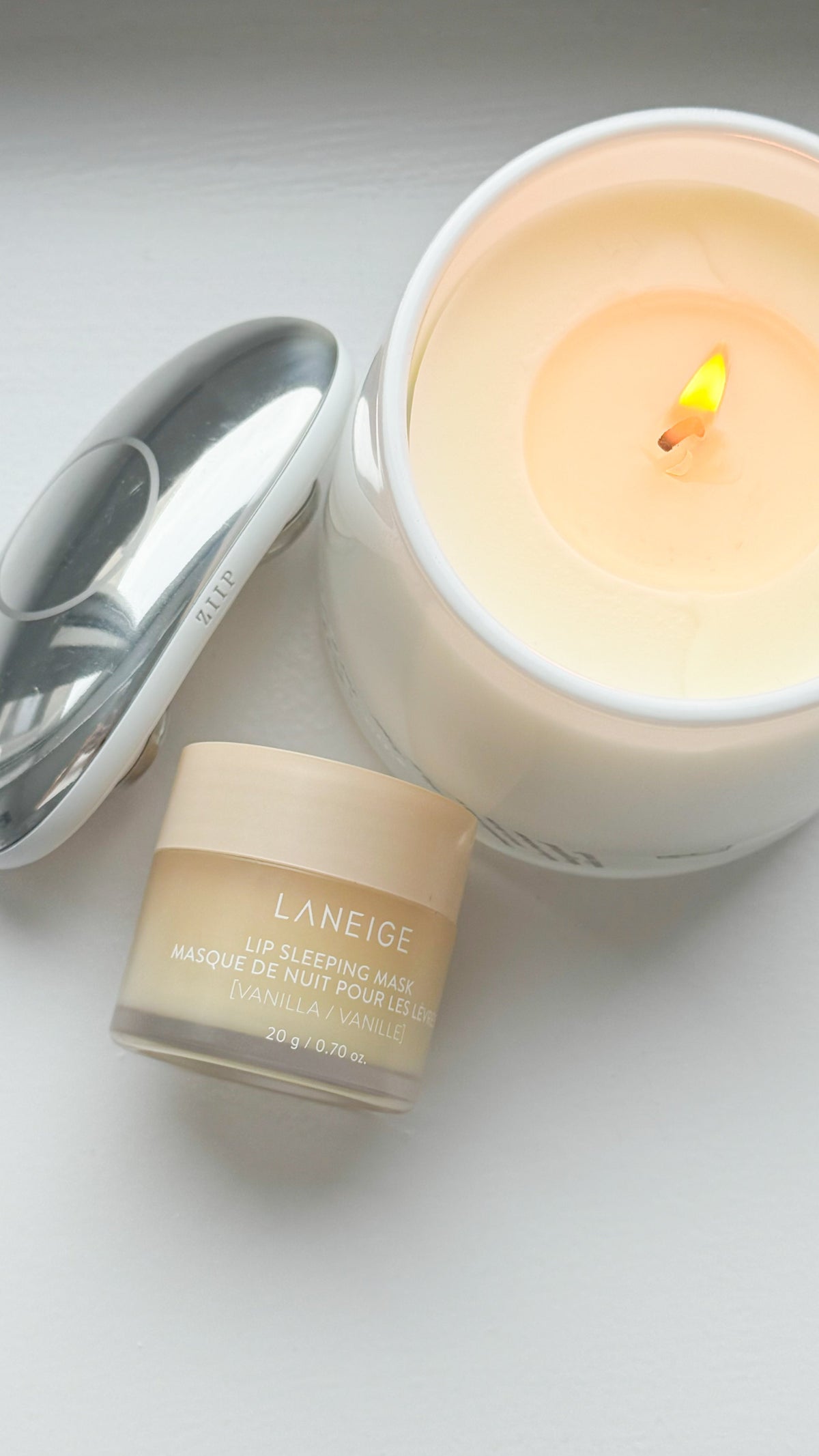 Flat-lay of a lit cream-colored candle, an open jar of Laneige Lip Sleeping Mask, and ZIIP HALO, styled together on a white surface.
